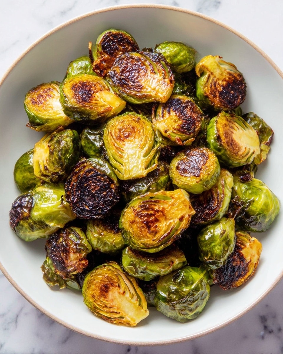 A white bowl filled with a pile of roasted Brussels sprouts, each cut in half. The Brussels sprouts have a mix of bright green and charred dark brown spots, showing a crispy, caramelized texture on the cut sides, while the outer leaves remain slightly wrinkled and deep green. The sprouts vary in size and are unevenly stacked in the bowl. The bowl sits on a surface with a white marbled texture. photo taken with an iphone --ar 4:5 --v 7