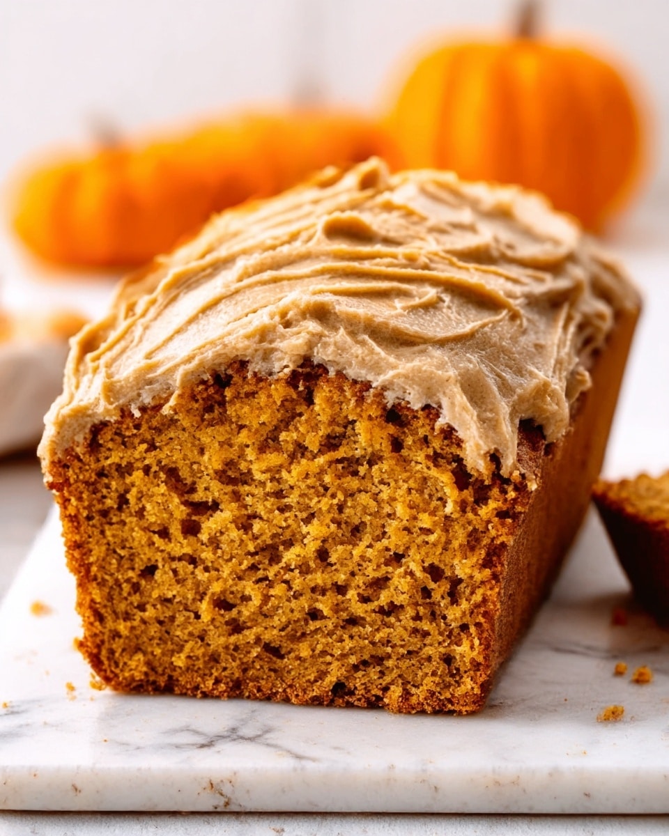 The image shows a loaf of pumpkin bread with a thick layer of light brown frosting on top that looks creamy and swirled. The bread inside is a warm orange-brown color with a moist, soft texture that is slightly crumbly. The loaf is cut to show the inside clearly, sitting on a white marbled surface with blurred oranges in the background. photo taken with an iphone --ar 4:5 --v 7