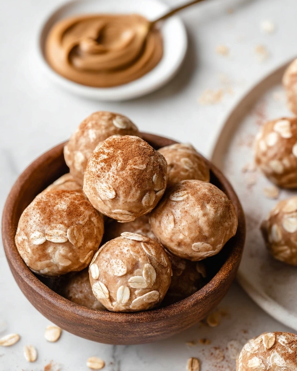 A close-up of a round brown bowl filled with light beige energy balls covered with large oat flakes and a dusting of cinnamon powder on top. The energy balls have a smooth, slightly textured surface with visible oats embedded throughout. Behind the bowl, a white plate holds a spoon with creamy peanut butter, smooth and light brown in color. In the bottom right corner, part of a white plate with more oat-covered energy balls is visible. The background is a white marbled texture with some scattered oat flakes and light brown powder. Photo taken with an iphone --ar 4:5 --v 7