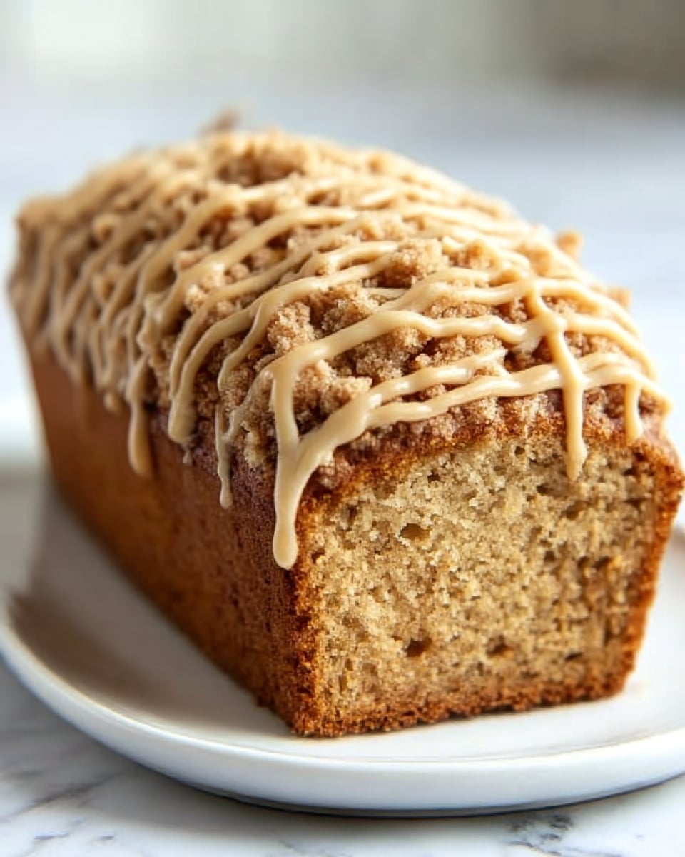 A loaf cake sits on a simple round white plate over a white marbled surface. The cake has a dense, golden-brown base layer with a thick crumbly topping made of streusel, which is light brown and rough in texture. Drizzled over the top is a pale beige glaze, flowing in thin, uneven lines adding a shiny finish. The cake's texture looks moist and soft underneath the crunchy topping. photo taken with an iphone --ar 4:5 --v 7