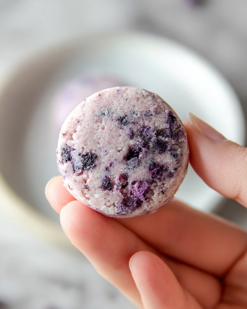 A close-up image of a small round cookie held between a woman's hand's fingers, showing a textured surface with a pale lavender color mixed with darker purple spots, suggesting crushed berries or similar ingredients inside. The cookie displays a rough, crumbly texture with visible unevenness and small chunks embedded throughout. In the blurred background, a white plate with another cookie is visible on a white marbled surface. photo taken with an iphone --ar 4:5 --v 7