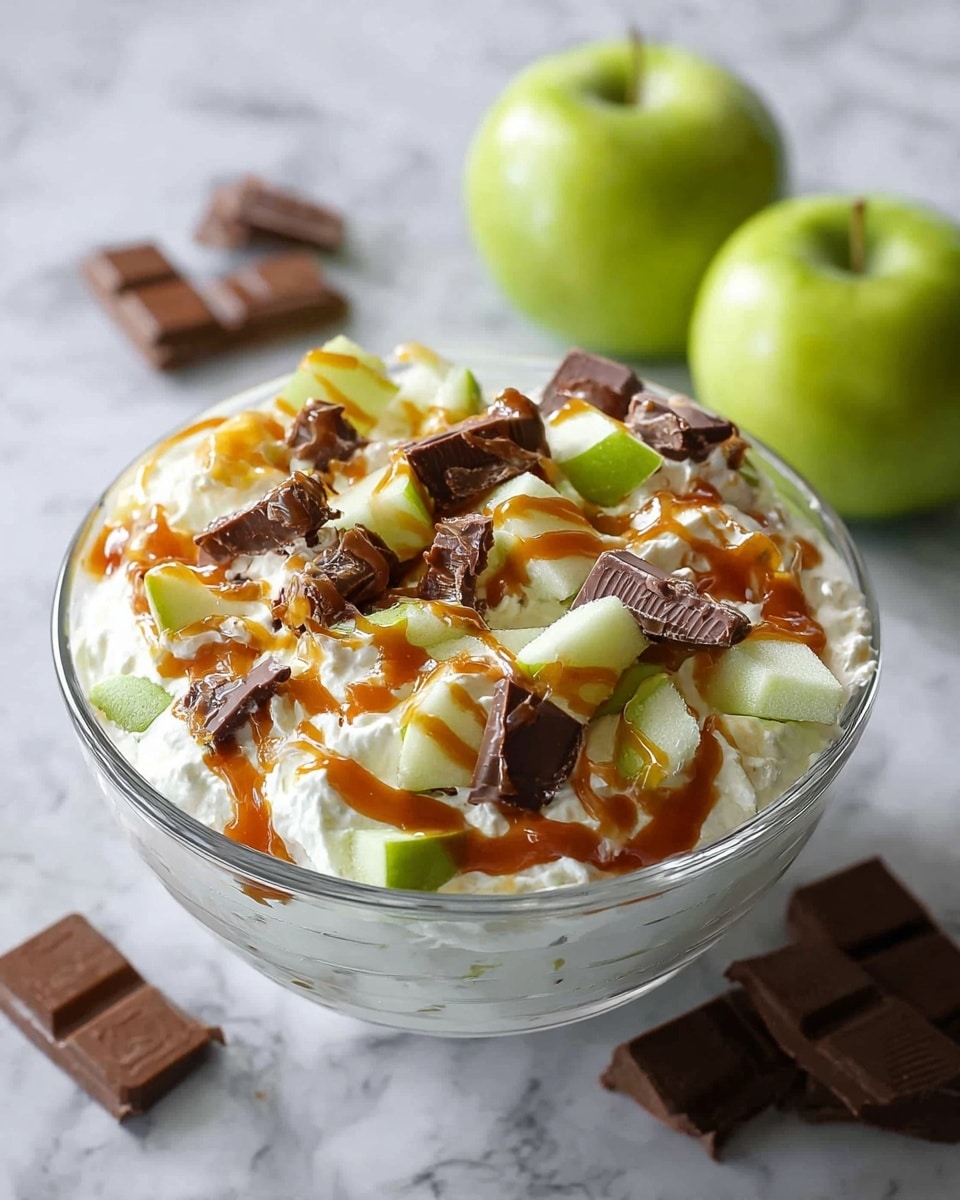A clear glass bowl is filled with a creamy white base, likely whipped or layered, covered with small pieces of green apple slices and chunks of chocolate candy scattered on top. A rich caramel sauce is drizzled over the top, adding an orange-brown glossy layer that contrasts with the white and green. The bowl sits on a white marbled surface with two whole green apples and some chocolate pieces placed in the background, giving a fresh and sweet atmosphere. photo taken with an iphone --ar 4:5 --v 7