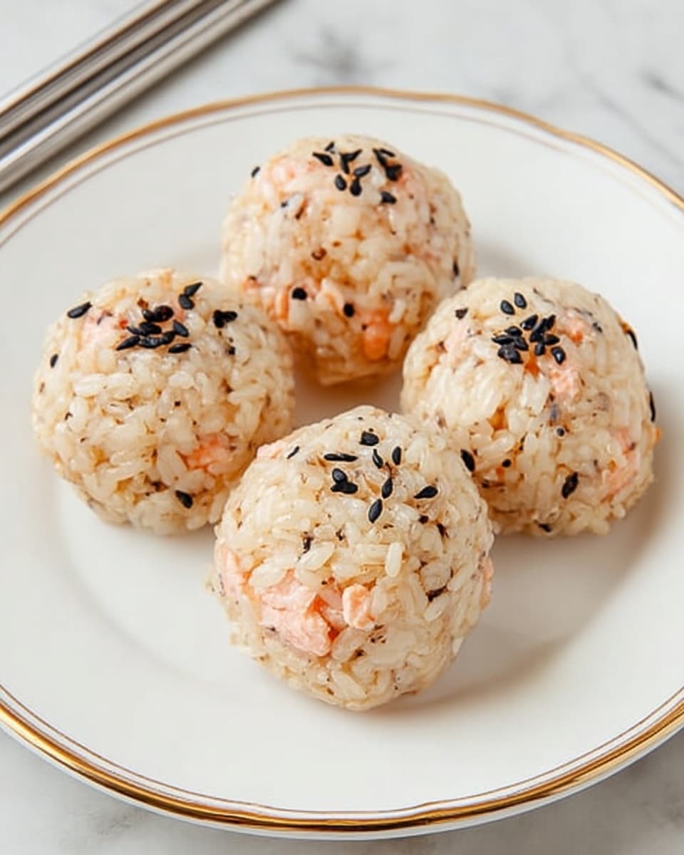 Four rice balls are placed closely together on a white plate with gold trim, each shaped into a round ball. The rice is light brown with visible small pieces of pink cooked salmon mixed throughout, creating a soft texture. On the top of each ball, there is a sprinkle of small black sesame seeds, adding a contrast in color. The plate is set on a white marbled surface with stainless steel chopsticks resting beside it. The overall look is clean and simple, focusing on the natural colors and textures of the rice and salmon. photo taken with an iphone --ar 4:5 --v 7