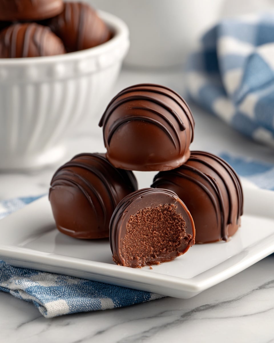 The image shows three round chocolate truffles on a white square plate placed on a white marbled surface. The truffles are covered in smooth, dark chocolate with thin drizzles of chocolate creating a striped pattern on the top and sides. One truffle is positioned upright, and the other two rest against it, with the front one partially bitten to reveal a creamy, dense, dark chocolate filling inside. In the background, a white bowl holds more truffles, and there is a blue and white checkered cloth nearby, blurred to keep focus on the truffles at the front. photo taken with an iphone --ar 4:5 --v 7