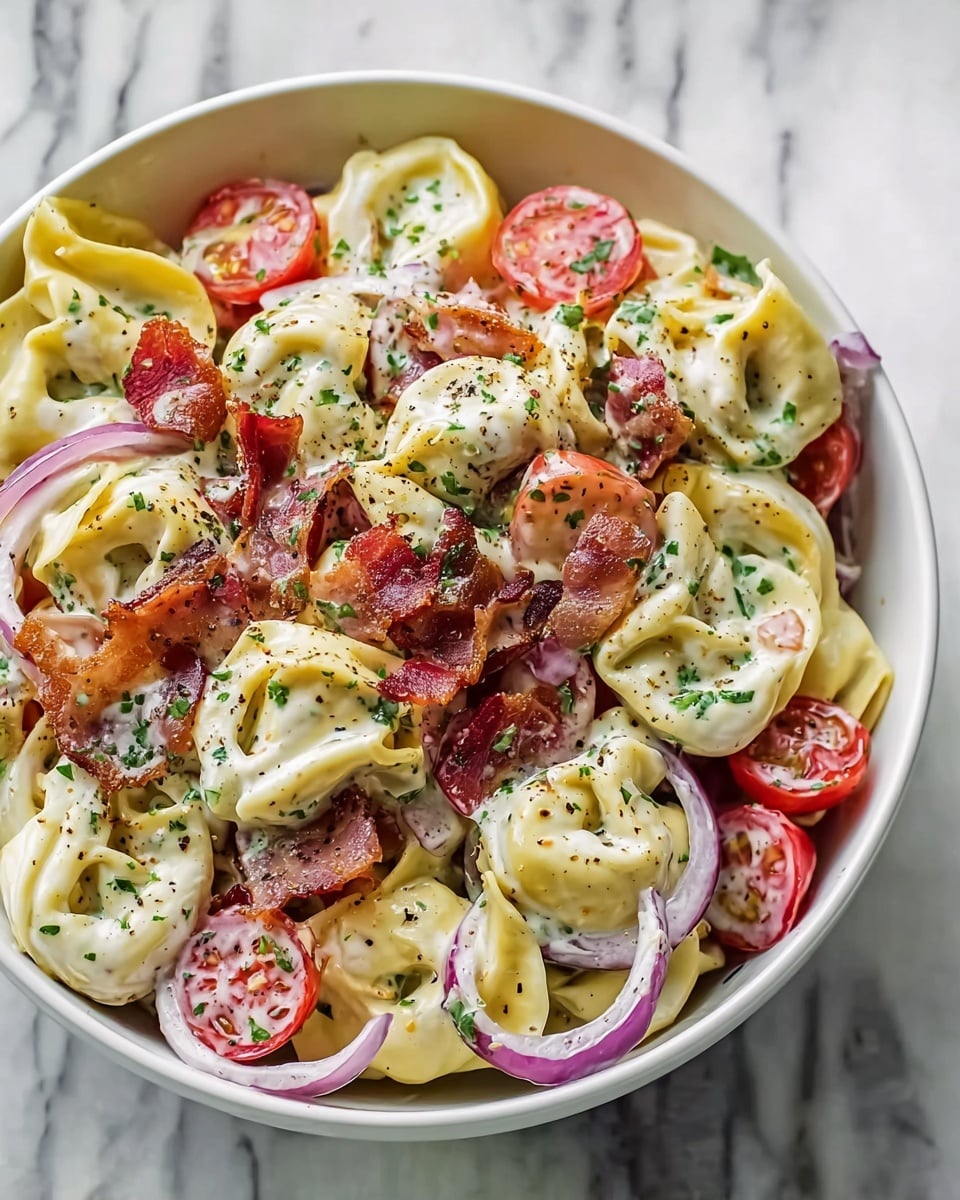 A white bowl filled with a creamy tortellini salad sits on a white marbled surface. The dish has several layers: the base is filled with pale yellow tortellini pasta, each piece smooth and plump, coated in a white creamy dressing. Mixed in are bright red cherry tomato halves and thin slices of purple-red onion rings. Scattered on top are crispy, dark reddish-brown bacon pieces and light green pepper slices, all sprinkled with finely chopped green herbs and black pepper. The colors and textures create a fresh and rich look. photo taken with an iphone --ar 4:5 --v 7