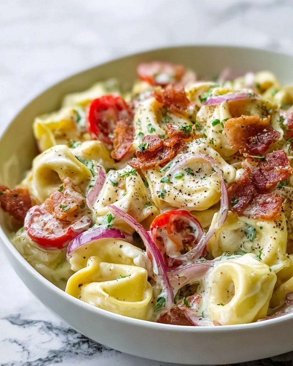 The dish is in a white bowl placed on a white marbled surface, showing a close view of creamy tortellini pasta. The pasta has pale yellow rings filled and folded, coated in a thick white creamy sauce. Mixed in are bright red cherry tomato halves, some glossy and shiny, along with thin rings of purple onions that add a soft texture. Scattered on top are small green herb pieces and bits of crispy reddish-brown bacon. A light sprinkle of black pepper dots the surface, giving a speckled effect. The overall presentation looks fresh and rich with colors and textures blending evenly. Photo taken with an iphone --ar 4:5 --v 7