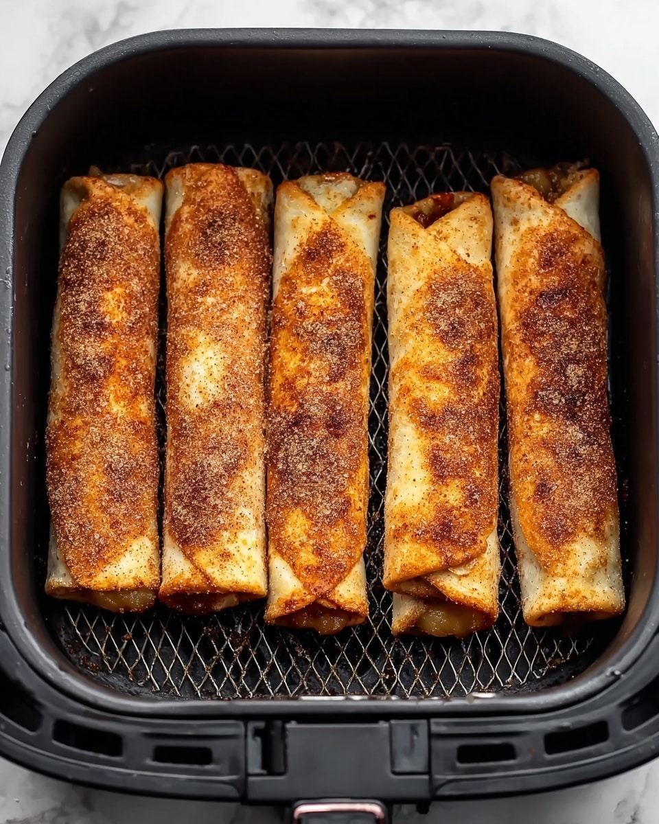 The image shows four rolled pastry sticks inside a black air fryer basket, lined up next to each other. Each roll is golden brown with a crispy texture and appears to be coated with a layer of cinnamon and sugar, giving them a slightly rough surface. The rolls show slight bubbling and caramelization spots, especially near the edges, while a few ends reveal a gooey filling that looks like baked apple or cinnamon filling peeking out. The air fryer basket has a fine mesh bottom and side walls, and the background is a white marbled texture. Photo taken with an iphone --ar 4:5 --v 7