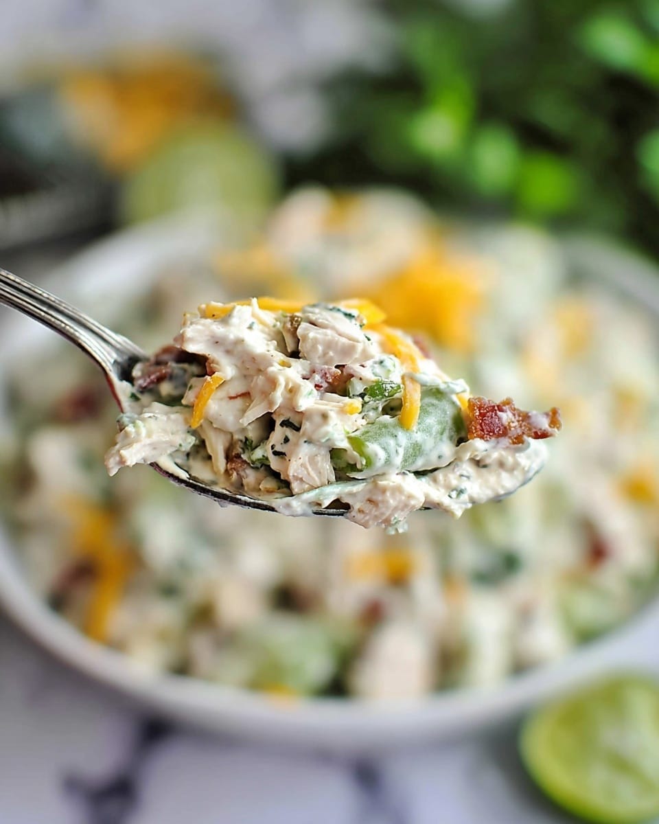 A close-up of a spoon holding a creamy salad mix with visible small chunks of white chicken, green celery pieces, shredded yellow and orange cheese, and small brown bacon bits, all blended in a thick white dressing. The background shows a white bowl filled with the same salad on a white marbled surface, with blurred green lime wedges at the bottom and leafy greens out of focus in the distance. The texture looks creamy and chunky, with a balanced mix of colors and elements. photo taken with an iphone --ar 4:5 --v 7