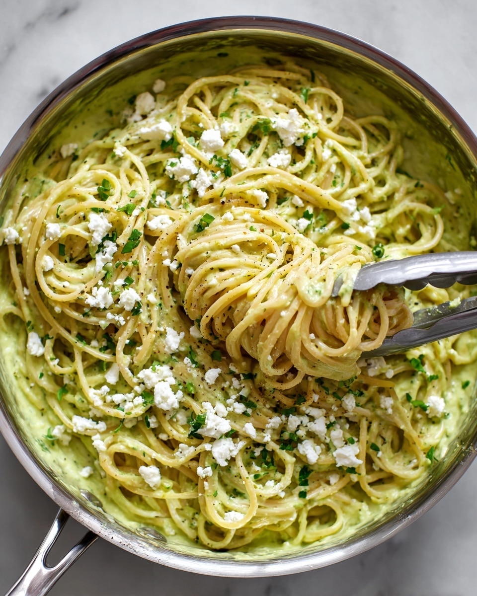 A close-up top view of a large silver pan filled with spaghetti pasta coated in a thick, creamy, light green sauce. The sauce looks smooth and covers the noodles evenly. White crumbly cheese is sprinkled on top, along with small pieces of chopped green herbs scattered across the surface. A pair of silver tongs is lifting a twist of pasta from the pan, showing the sauce clinging to the noodles. The scene is set against a white marbled surface. photo taken with an iphone --ar 4:5 --v 7