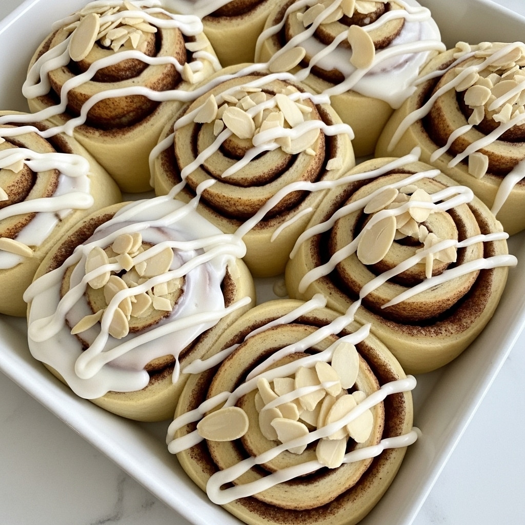 This close-up image shows several cinnamon rolls arranged closely in a white baking tray, each roll having a light golden-brown color with visible spiral layers of dough and cinnamon filling. On top, there is a thick layer of shiny white icing spread unevenly over the rolls, with extra icing drizzled in thin swirls that partly cover the spiral shapes. Scattered across the icing are thin, pale beige almond slices with slightly darker edges, adding texture and contrast. The tray is set against a white marbled surface that provides a clean and bright background. Photo taken with an iphone --ar 4:5 --v 7