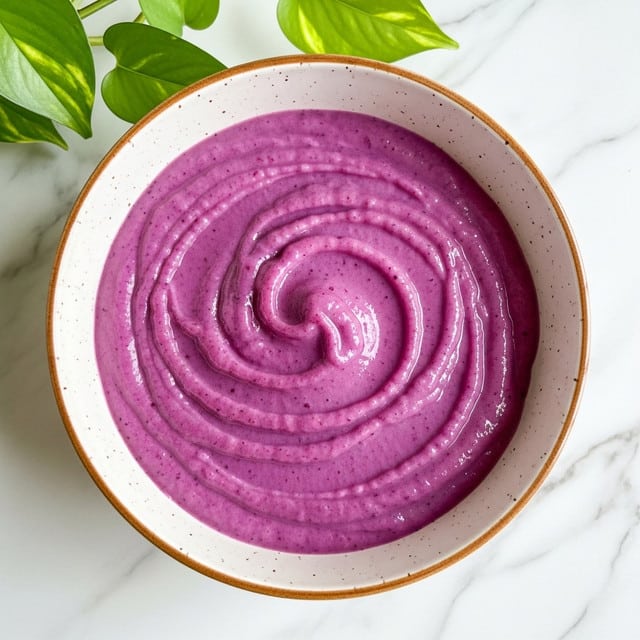 The image shows a bowl filled with a smooth, bright purple puree that has a creamy texture with gentle swirls on the surface, creating a slight spiral pattern. The bowl is white with subtle brown speckles, sitting on a white marbled surface. There is a green leafy plant partially visible at the top edge of the image, adding a fresh touch to the simple composition. photo taken with an iphone --ar 4:5 --v 7