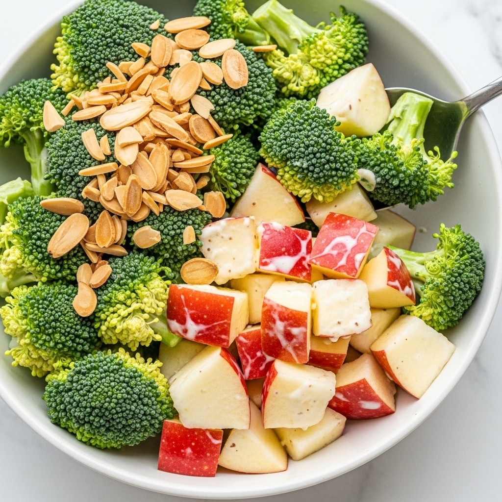 A close-up view of a broccoli salad showing roughly three layers: the first layer is fresh green broccoli florets with a bumpy texture, the second layer includes chunks of red apple with white flesh and smooth skin sitting lowered among the broccoli, and the third layer scattered on top consists of light brown sliced almonds with a dry, thin texture. The salad is lightly coated with a creamy white dressing, giving a slight shine to the broccoli and apple. The dish is in a white bowl on a white marbled surface with a silver spoon lifting a mix of broccoli and apple pieces. Photo taken with an iphone --ar 4:5 --v 7